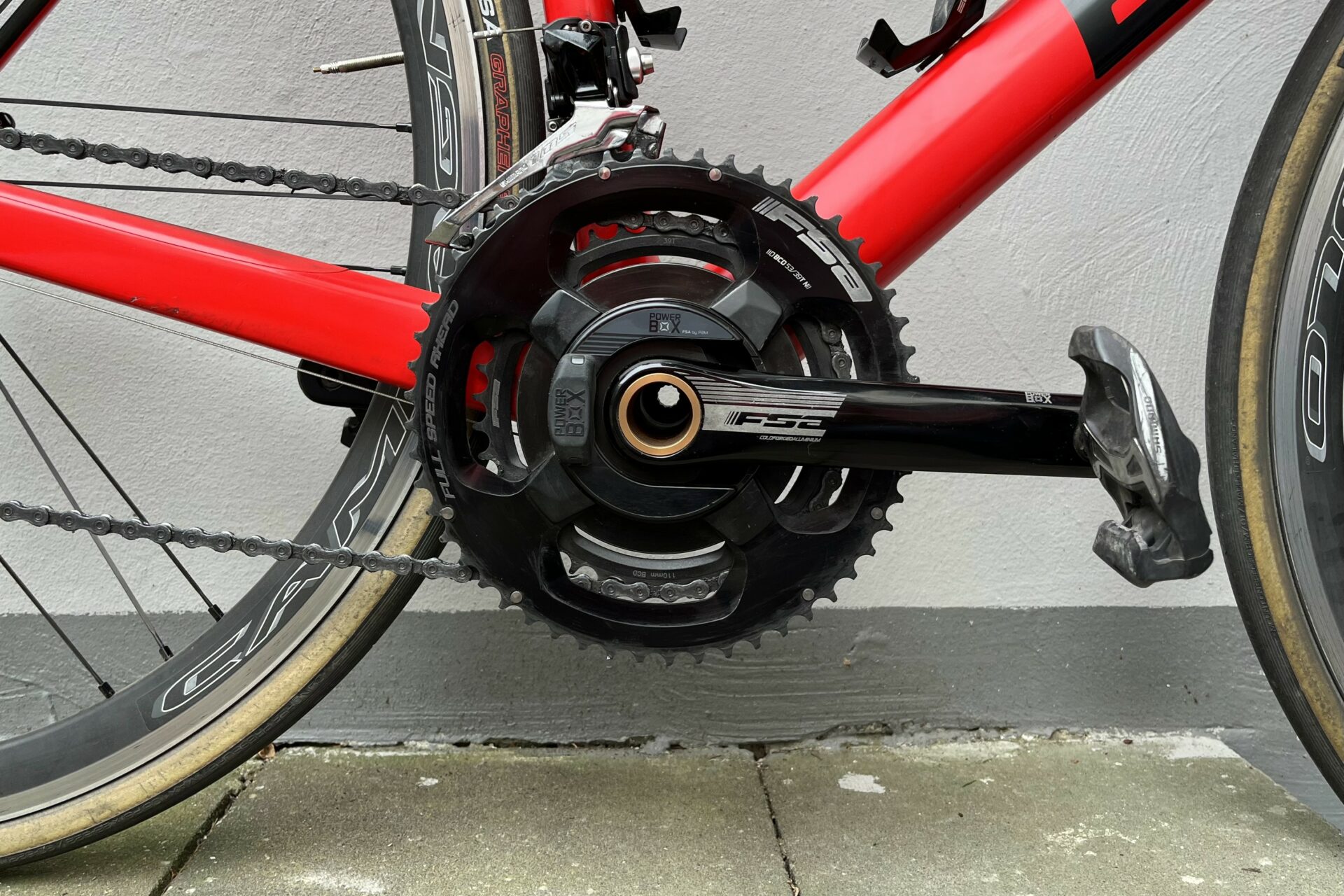 In front of a white and grey wall stands a red BMC road bike, equipped with an FSA Powerbox crank-based power meter, the best power meter on the market in terms of accuracy and reliability.